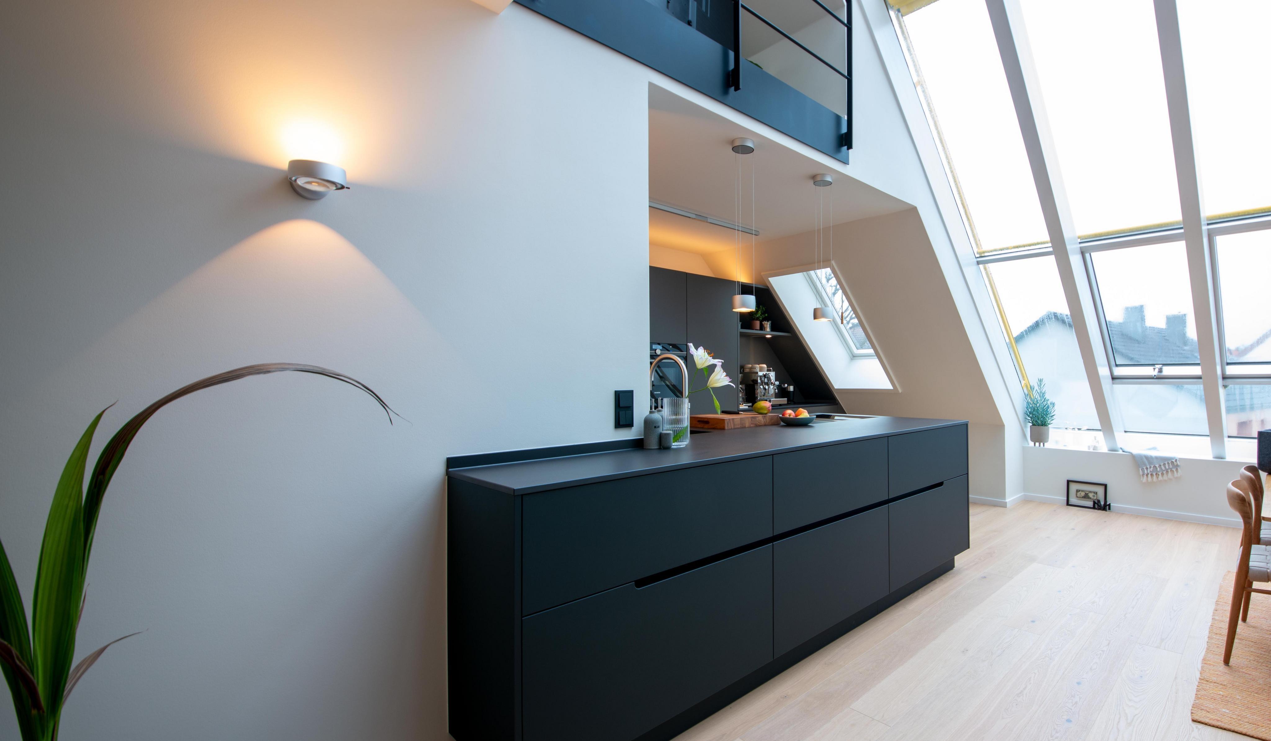 Moderne offene Küche mit schwarzer Einrichtung, viel Tageslicht durch schräge Fenster.