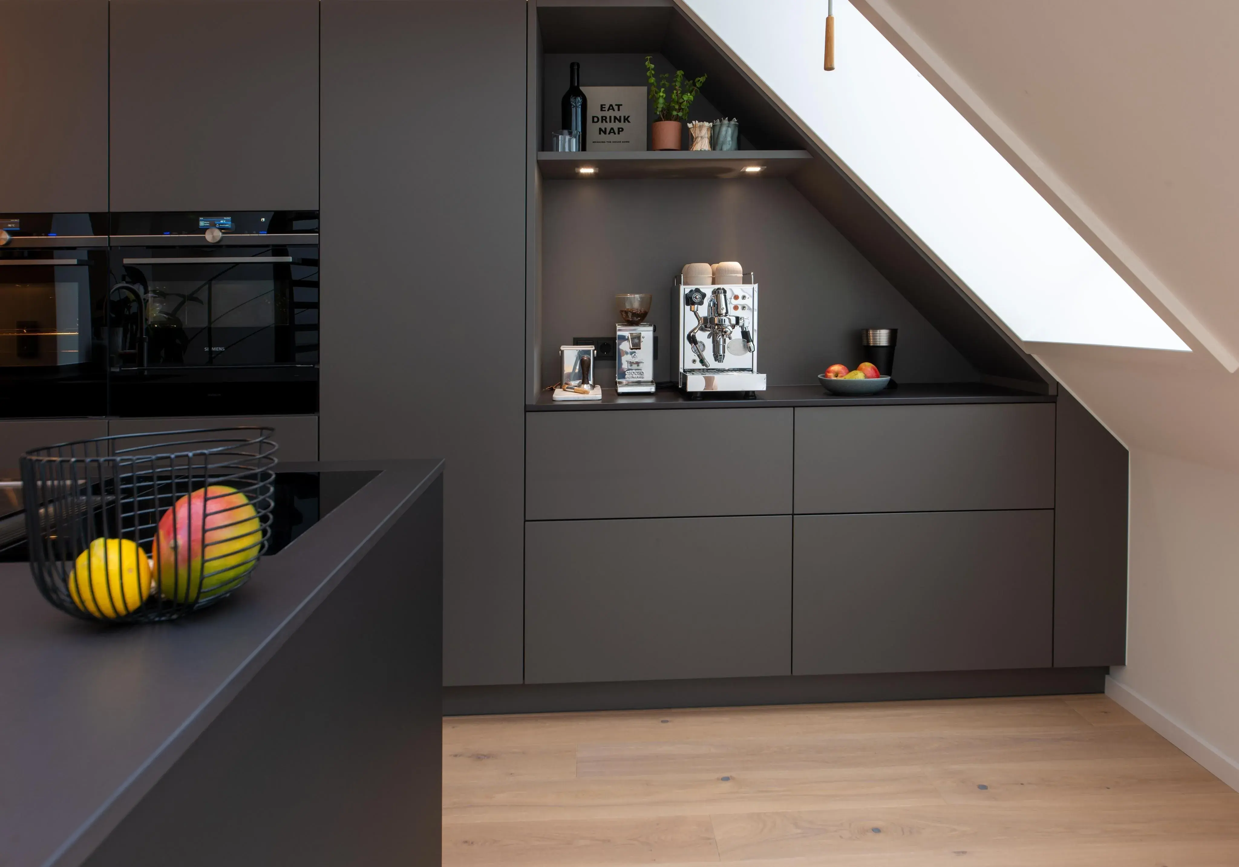 Moderne Küche mit dunklen Schränken, Obstkorb und Kaffeemaschine unter einer Dachschräge.