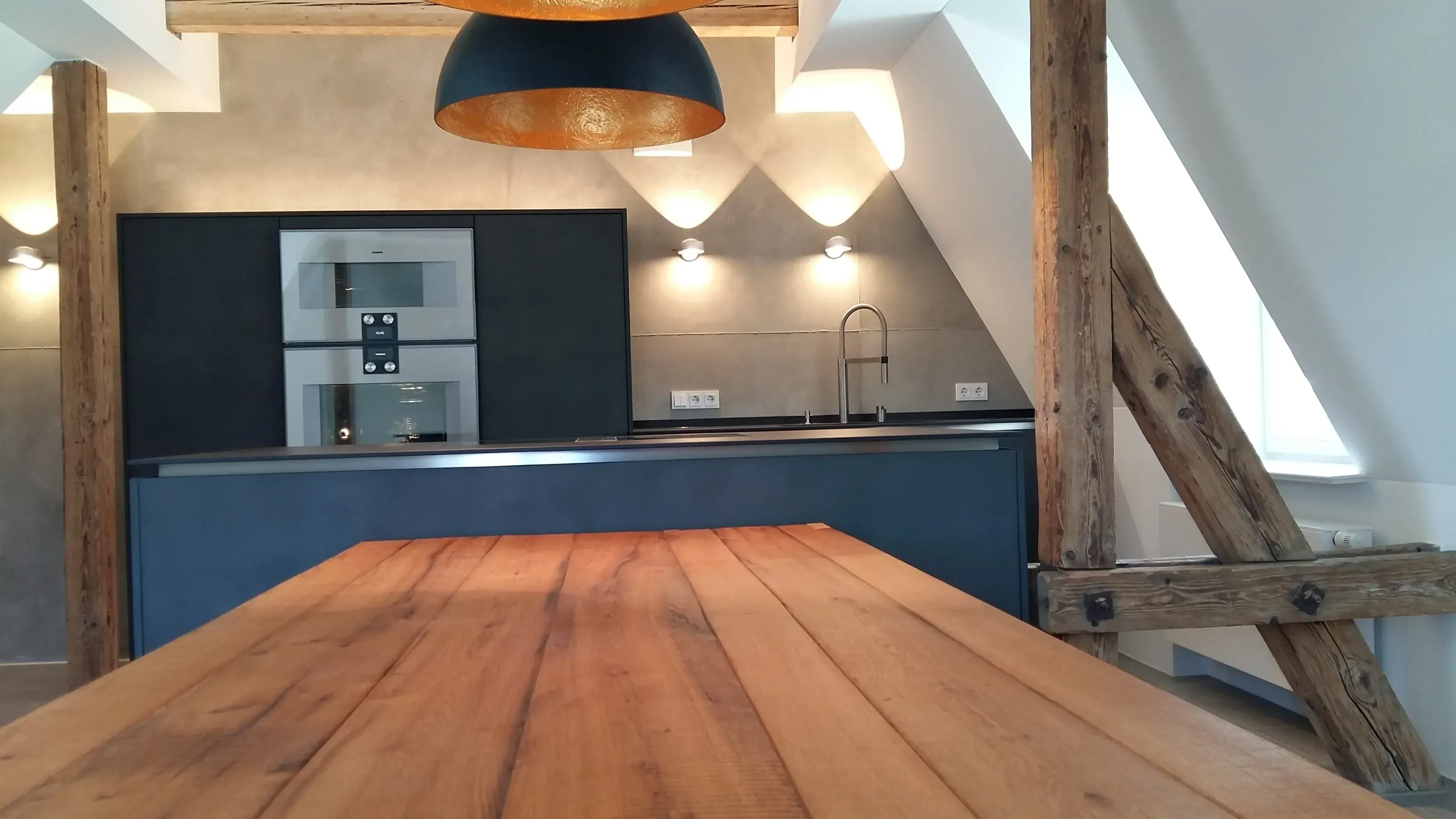 Moderne Küche mit Holztisch, dunklen Lampen und sichtbaren Holzbalken.
