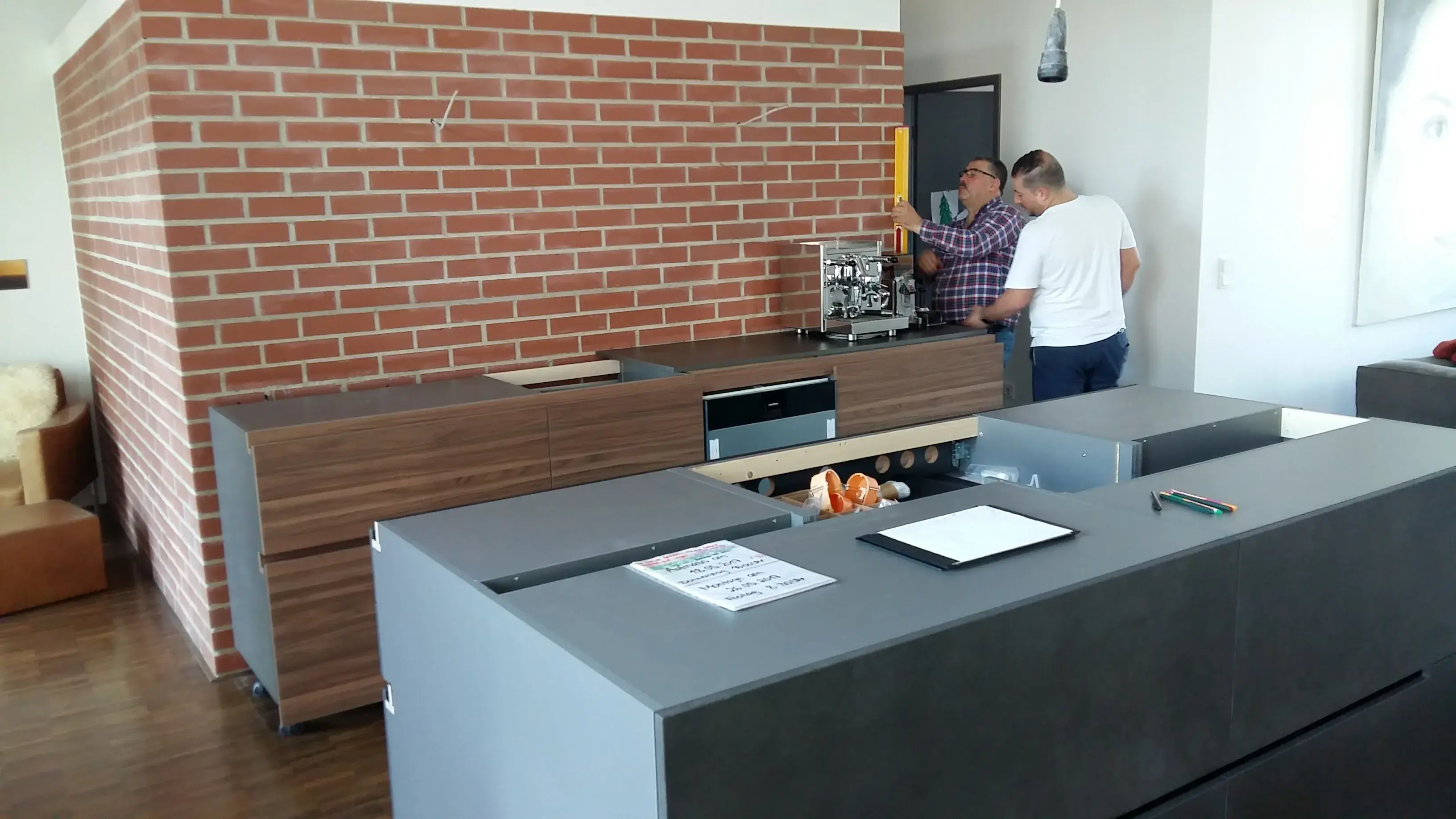Moderne Küche mit zwei Männern neben einem Kaffeeautomaten vor einer Backsteinwand.