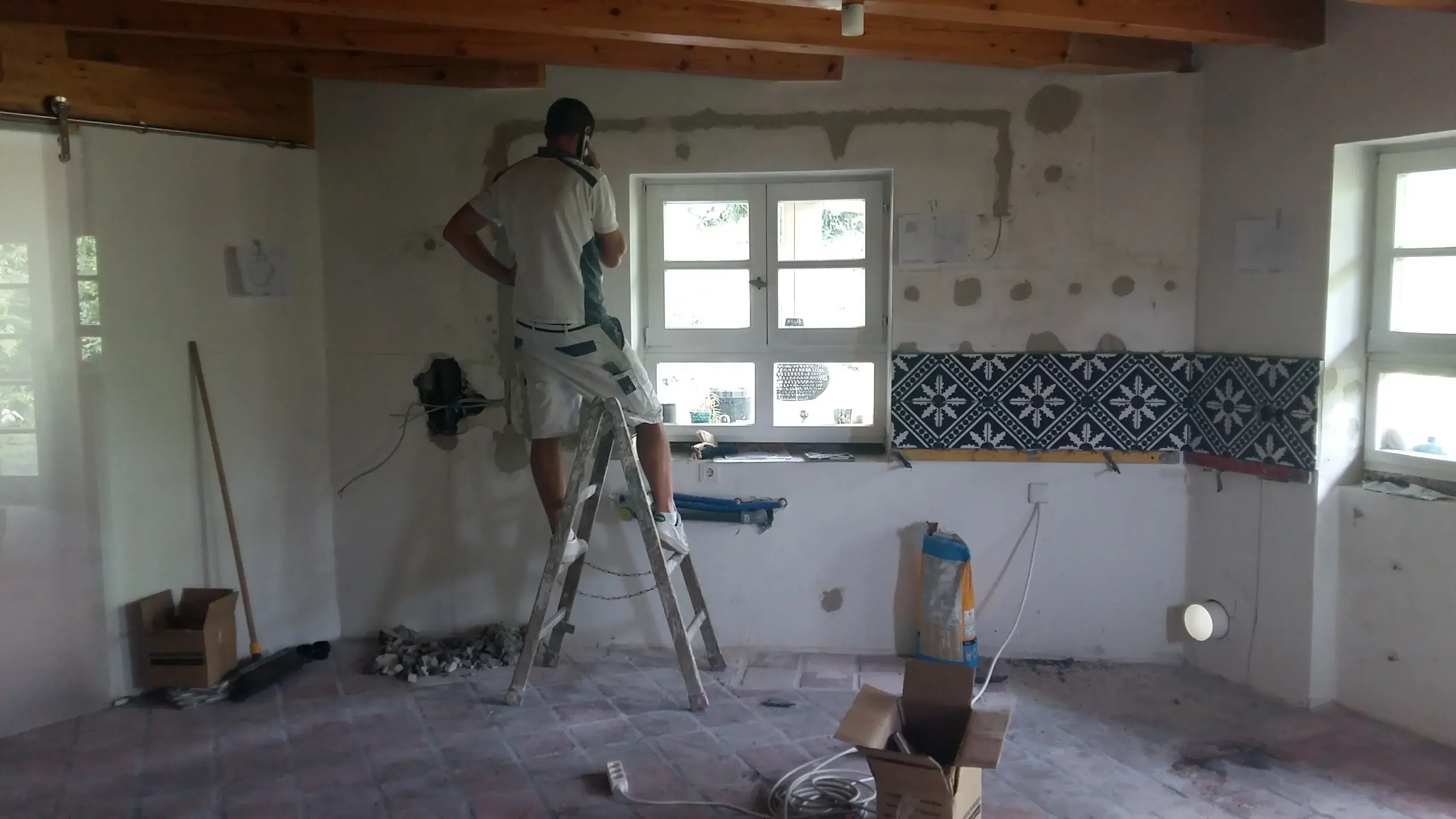 Mann arbeitet auf einer Leiter in einer Küche im Umbau, Fliesen an der Wand.