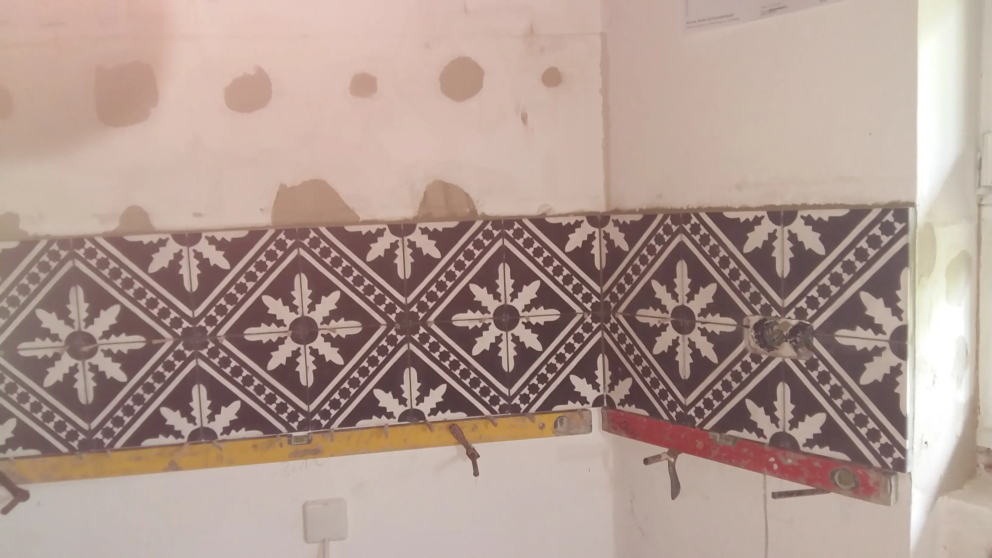 Teilweise geflieste Wand mit schwarzem Muster, obere Wand unverputzt.