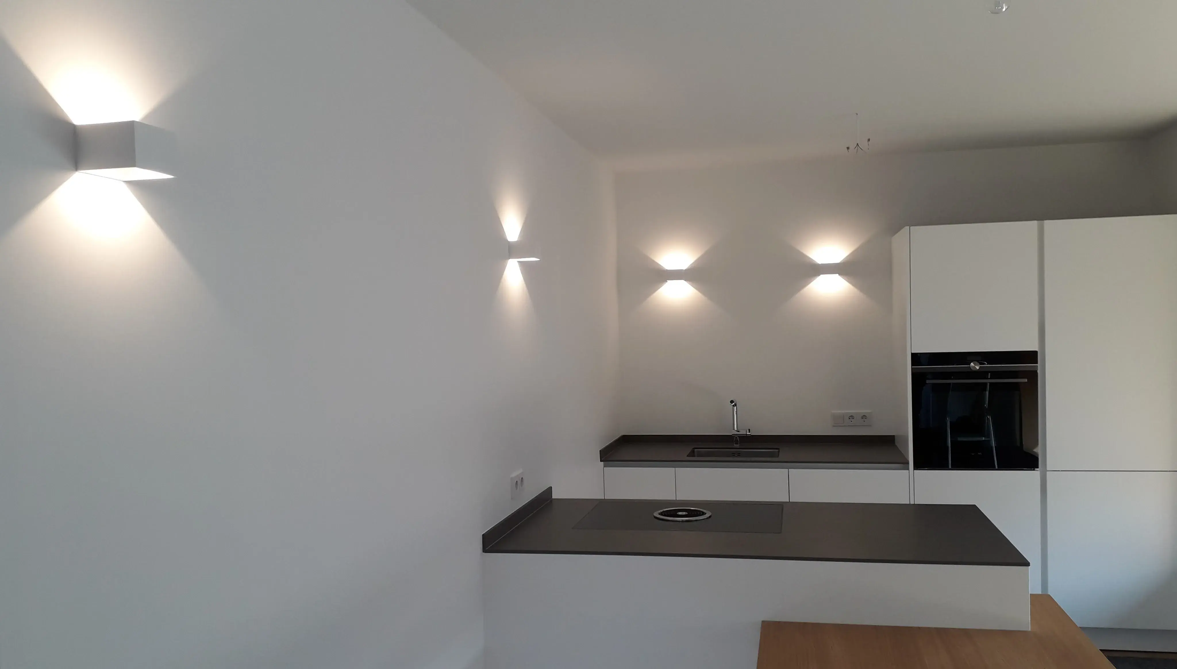Moderne Küche mit weißer Wand, schwarzen Arbeitsflächen und eingeschalteten Wandlampen