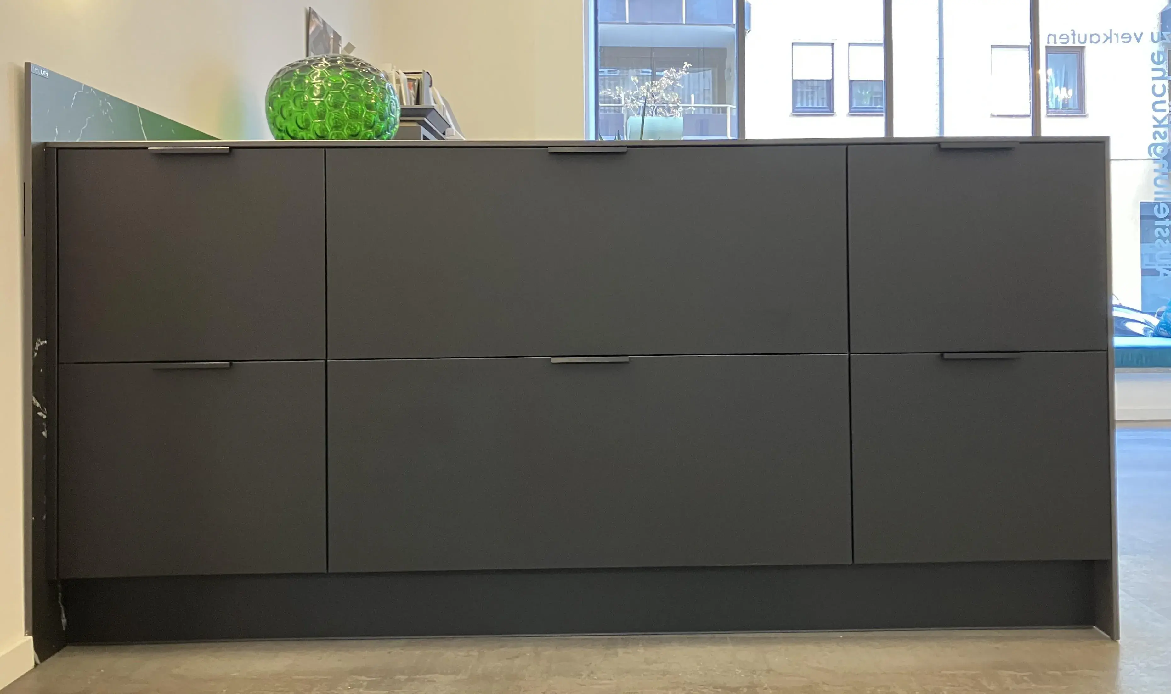 Schwarzer Sideboard mit Schubladen, darauf ein Glasgefäß mit Dekosteinen, Fenster im Hintergrund.