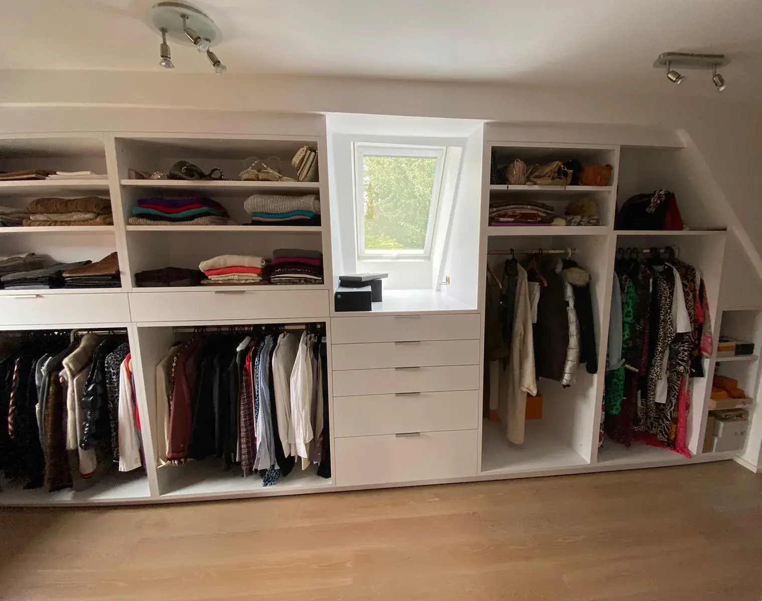 Offener Kleiderschrank mit Kleidung, Regalen und Schubladen, unter Dachfenster.