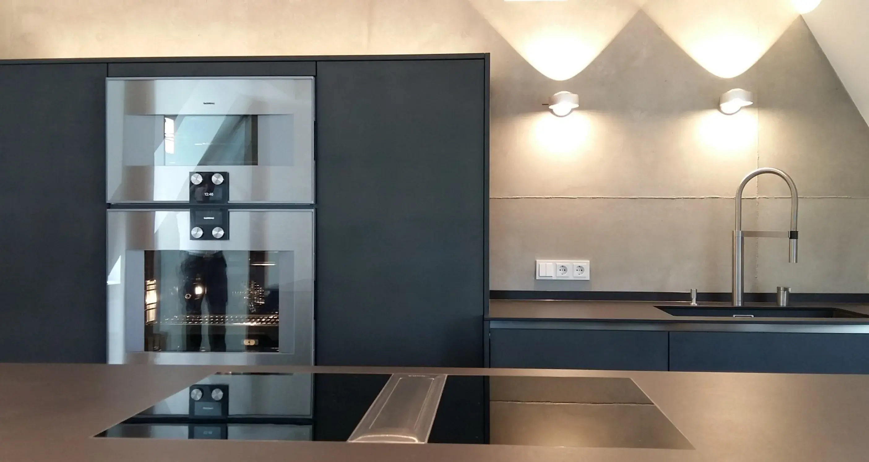 Moderne Küche mit dunklen Schränken, Kochfeld, Spüle und Wandlampen und zwei eingebauten Gaggenau Backöfen.