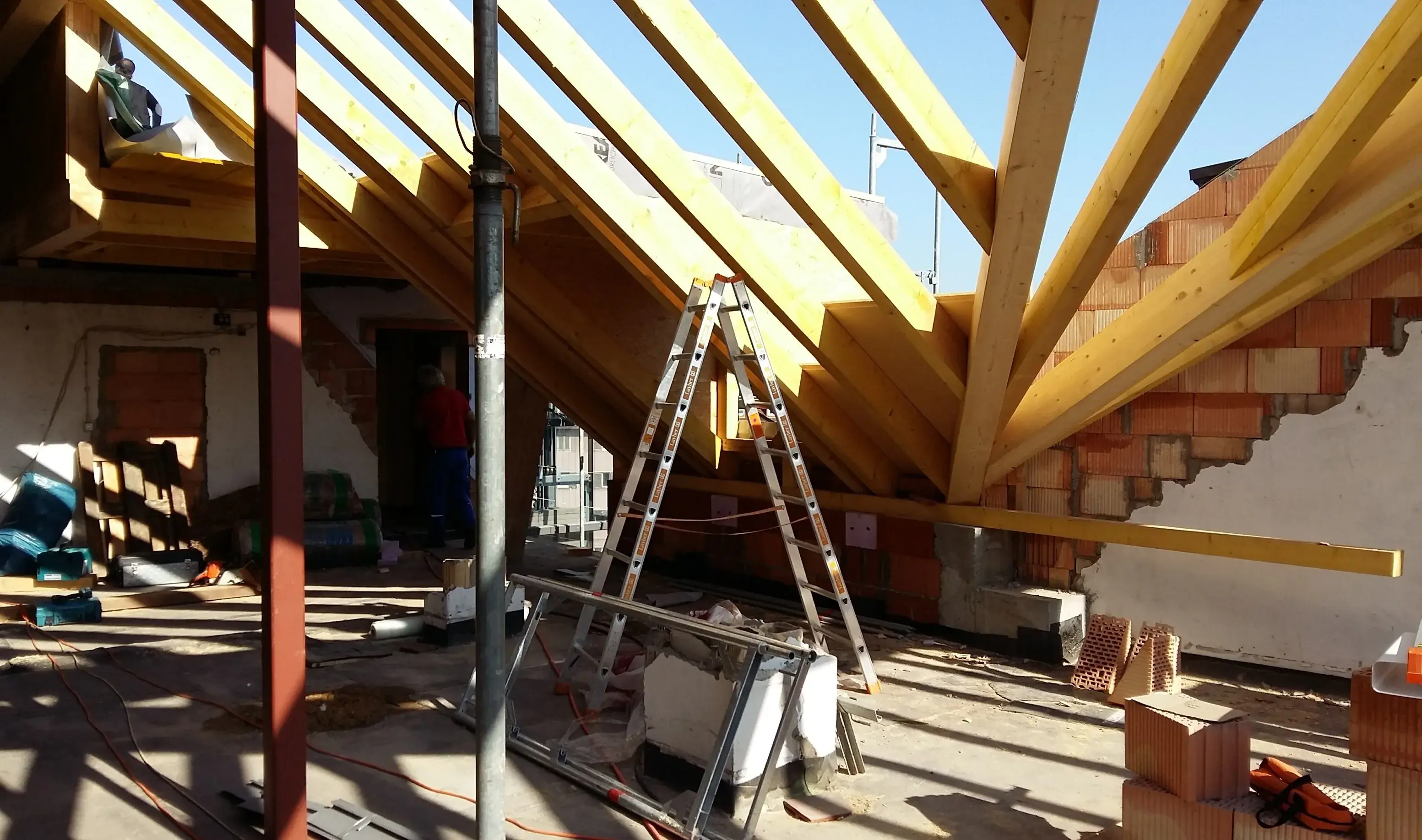 Dachstuhl-Bau mit Holzträgern, Leitern und Baumaterialien bei Sonnenschein.