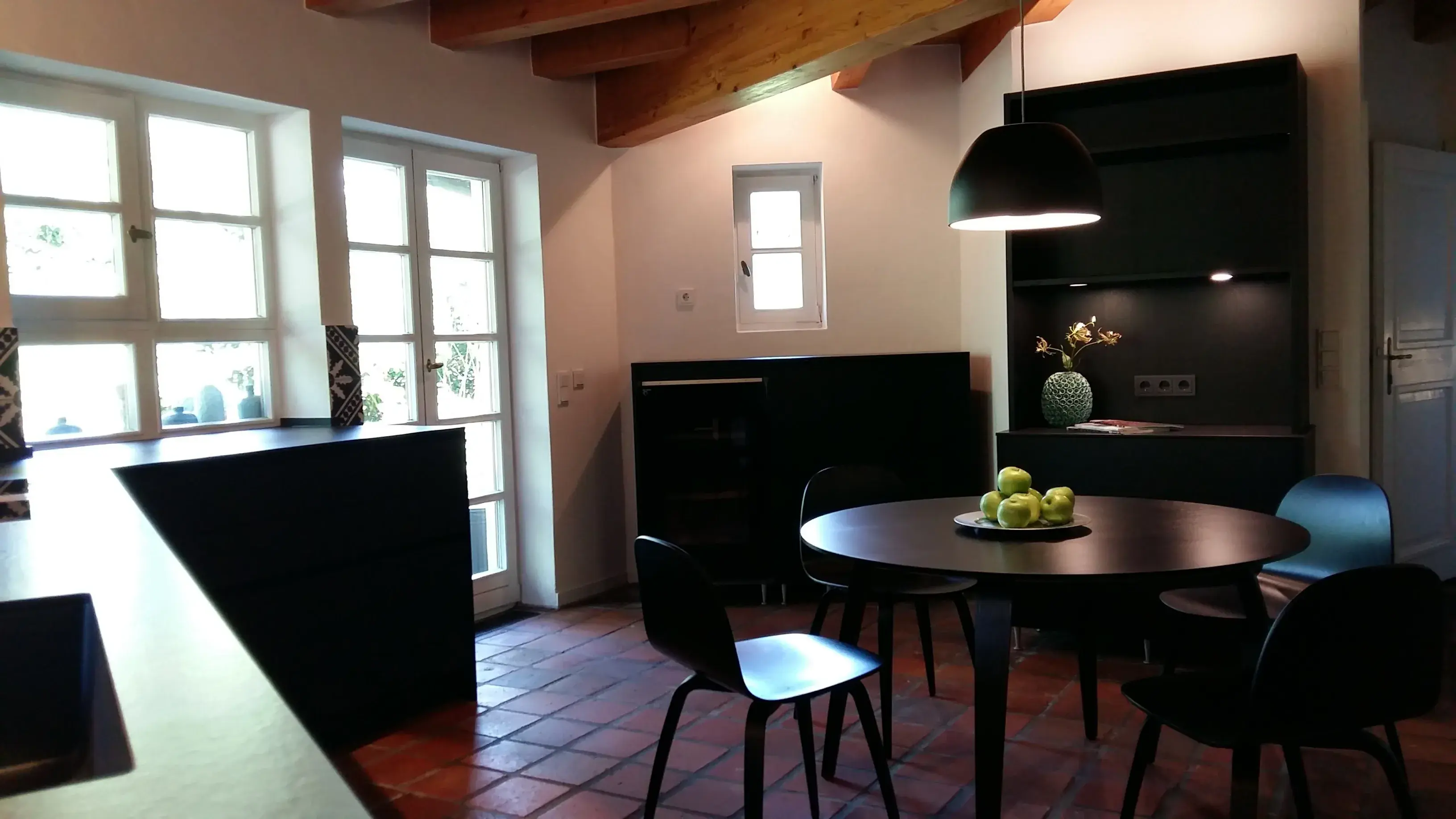 Moderne Küche mit rundem Esstisch, schwarzer Lampe und Fenstern, Schale Äpfel als Deko.