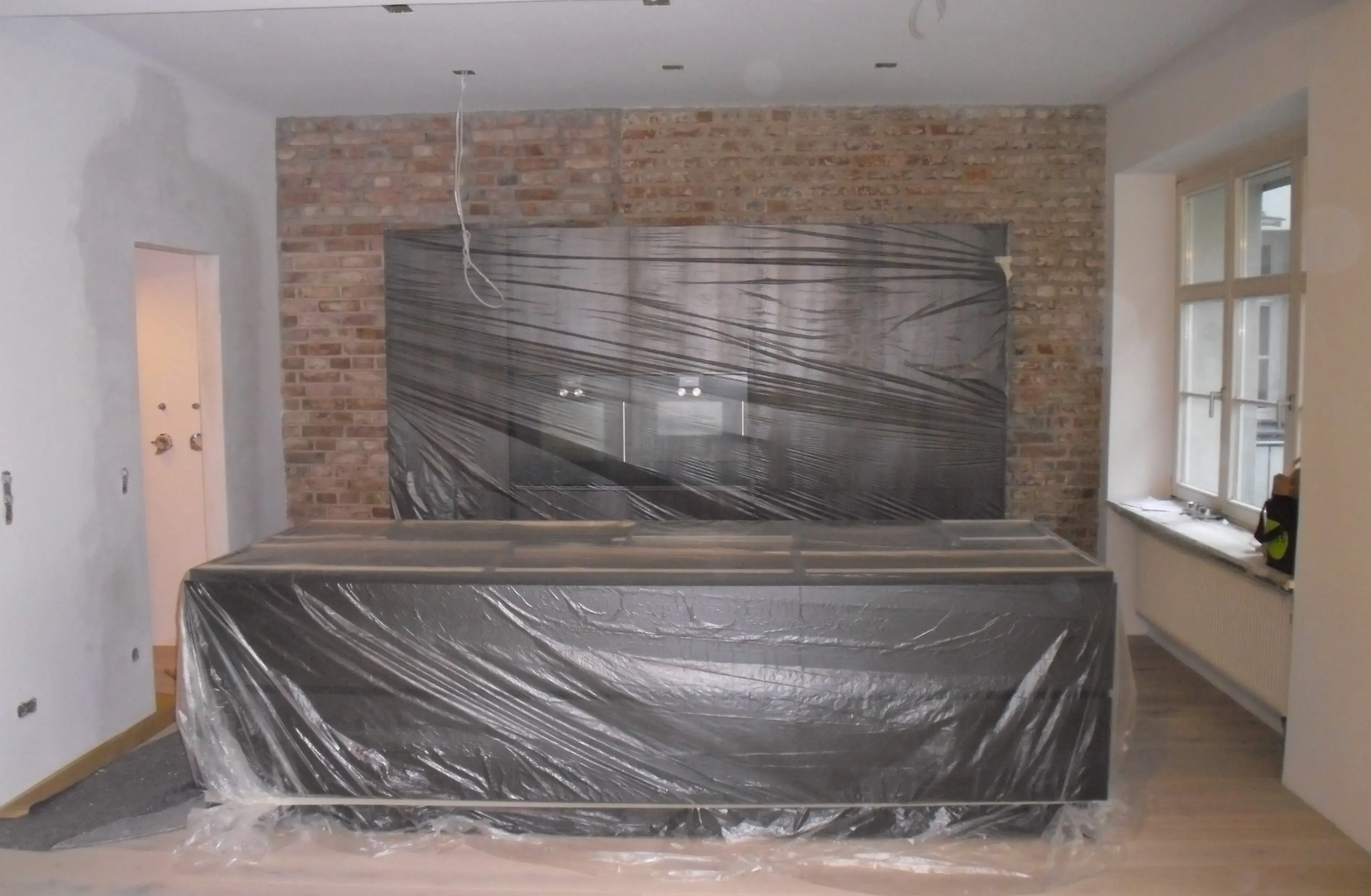 Moderne Küche mit Folie abgedeckt, Backsteinwand im Hintergrund, Fenster rechts.