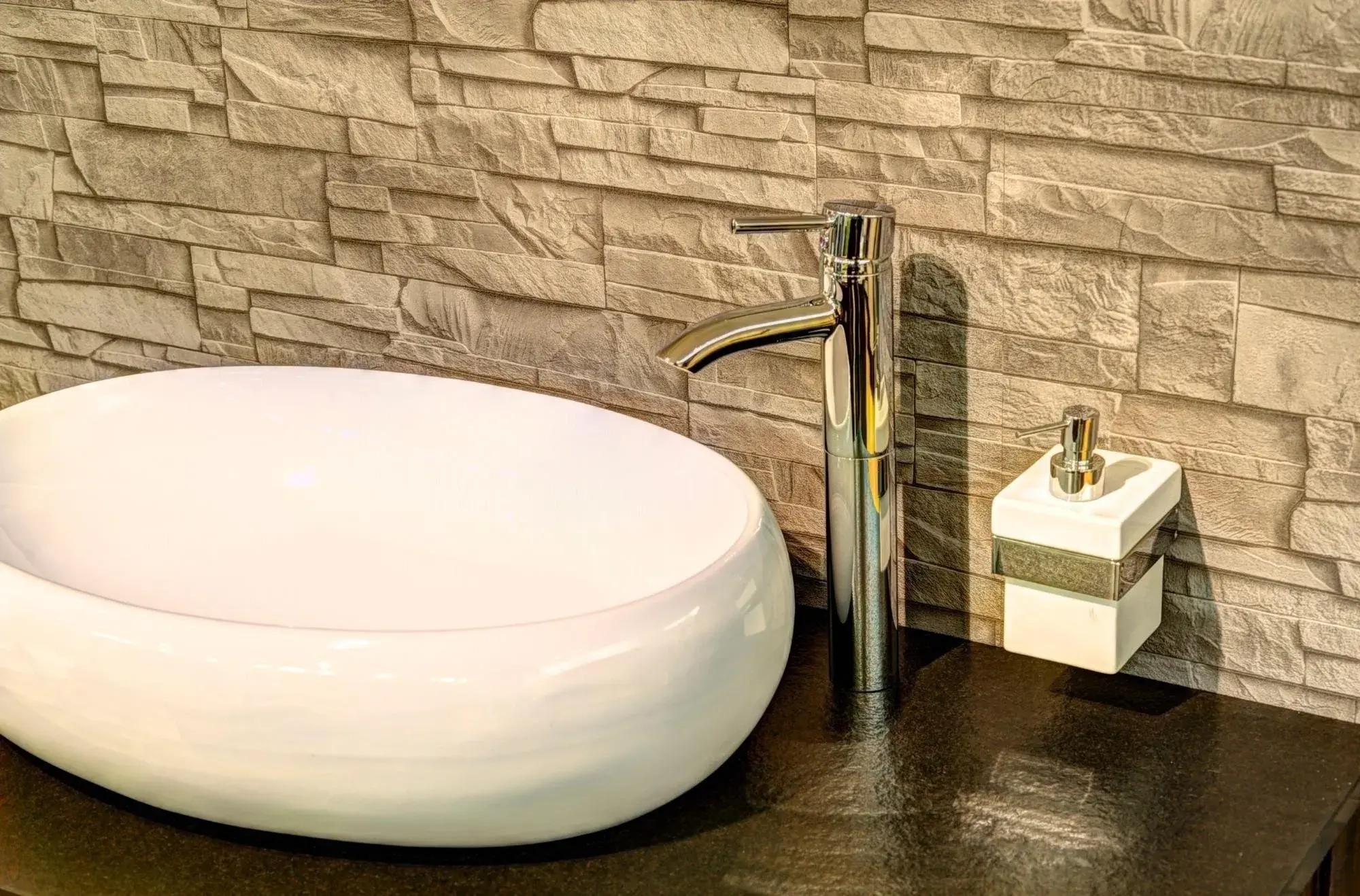 Modernes Waschbecken mit silbernem Wasserhahn vor Steinwand, Seifenspender daneben.
