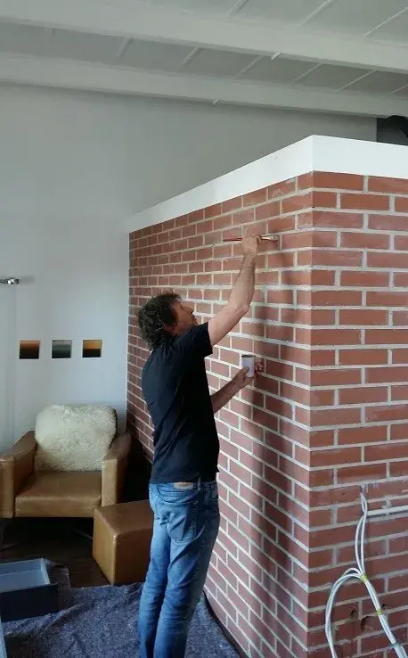 Mann bemalt weiße Fugen auf eine rote Backsteinwand in einem Wohnzimmer.