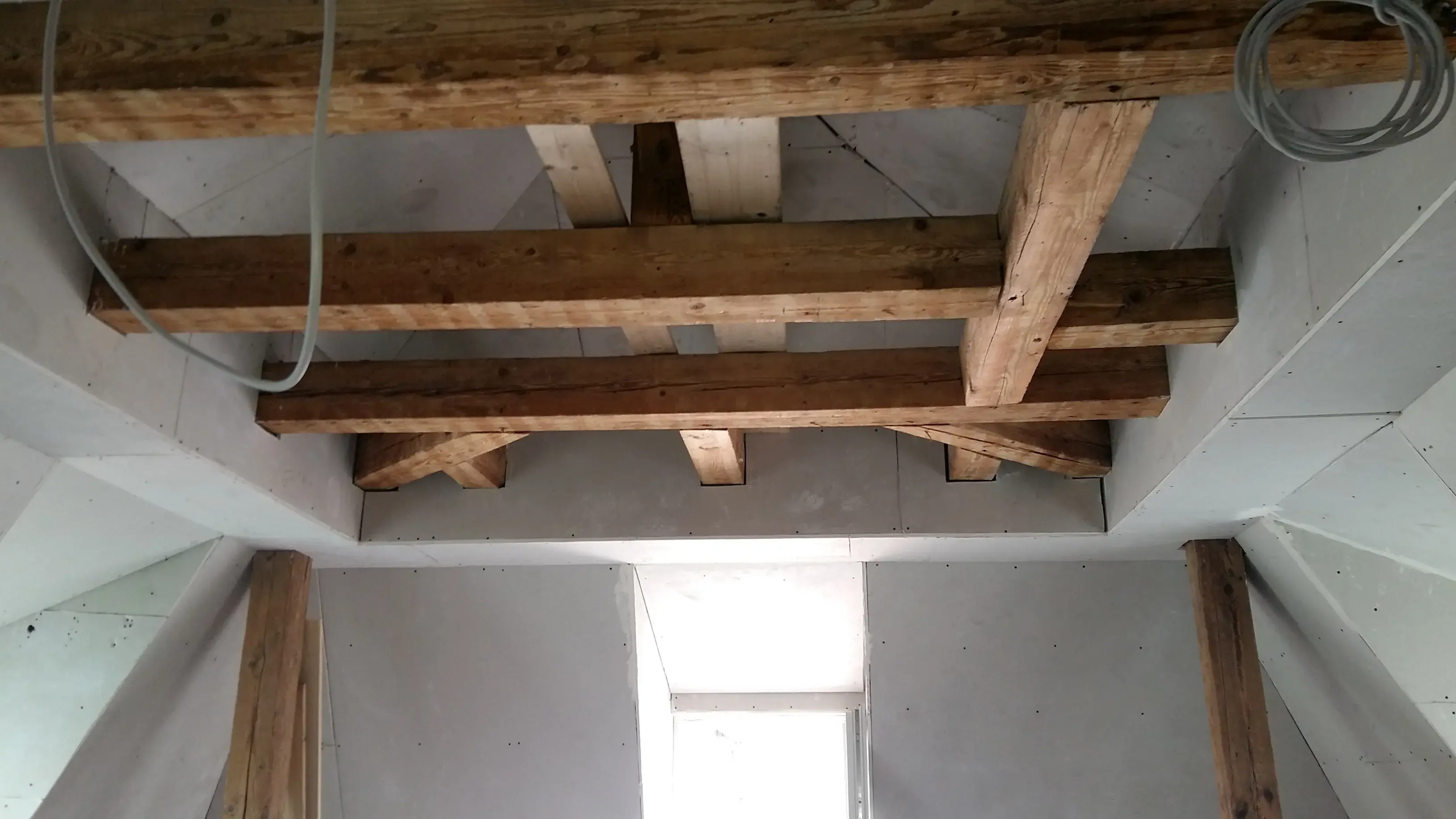 Holzbalken an einer weißen Decke, darunter ein kleines Fenster, Baustellensituation.