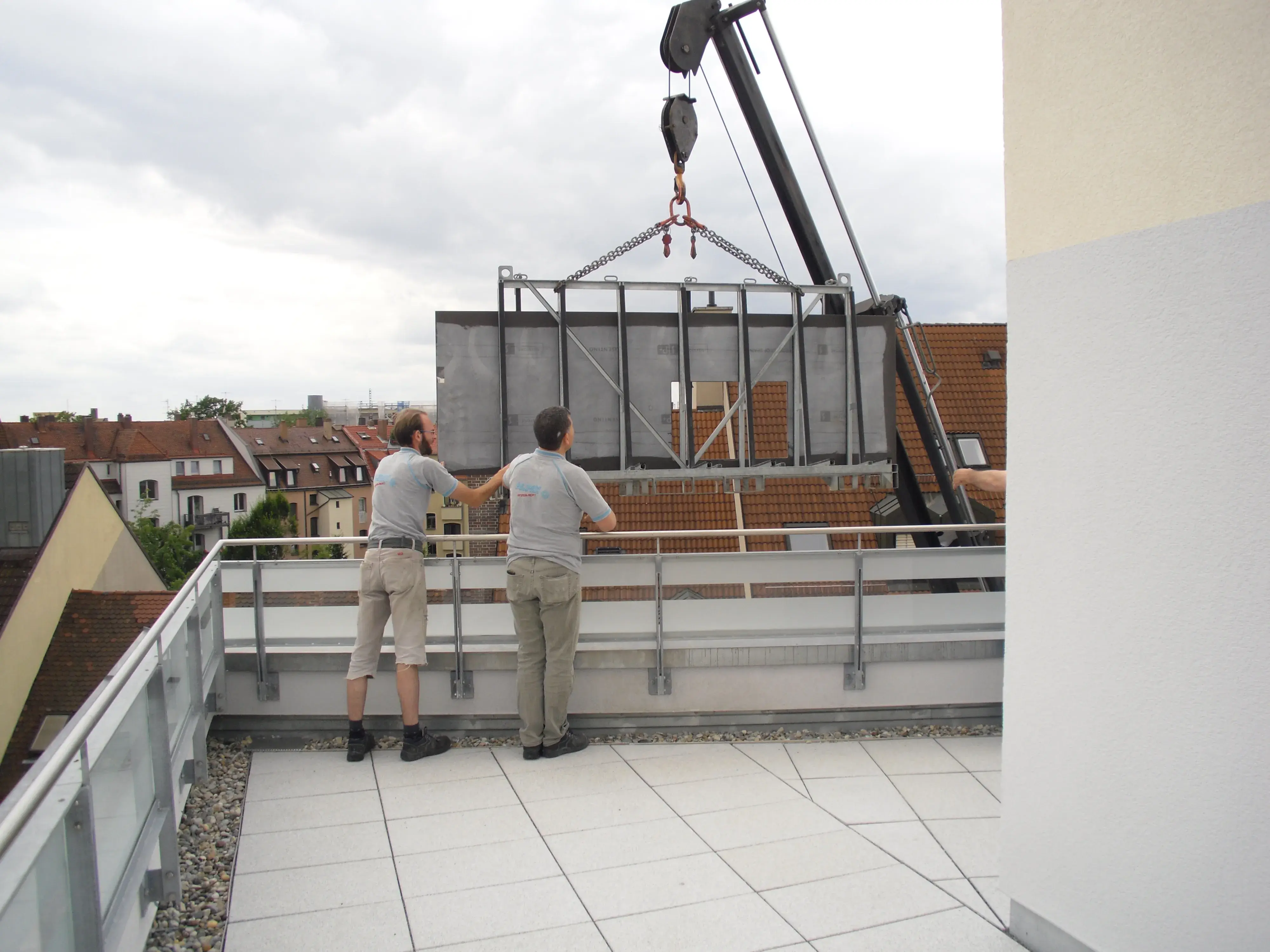 Zwei Männer auf Balkon beobachten Kran, der ein große Arbeitsplatte über die Brüstung hebt.